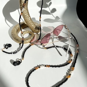 May include: Black beaded glasses chain with brown and white beads, and gold accents. The chain is draped over a gold snake statue and a pair of pink sunglasses. The chain has the words "Bite Me" spelled out in black beads.
