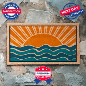 May include: A brown coir doormat with a graphic design of a stylized orange sun setting over blue and teal waves. The doormat has a black border.