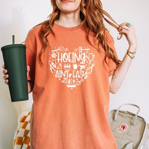 Op de afbeelding: Oranje T-shirt met een witte hartvormige afbeelding met de tekst "HOEING AIN'T EASY" met tuingereedschap en planten. Een donkergroene getextureerde beker wordt in de linkerhand gehouden. Het shirt is een comfortabele, casual stijl.