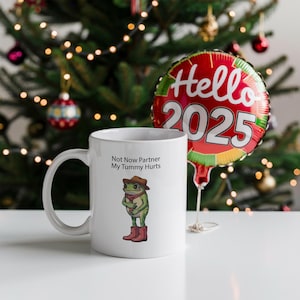 May include: White ceramic mug with the text "Not Now Partner My Tummy Hurts" and a cartoon frog wearing a cowboy hat and boots. A red and gold balloon with "Hello 2025" is in the background.