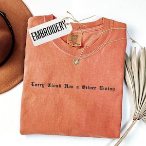 May include: A coral-colored t-shirt with the phrase "Every Cloud Has a Silver Lining" in black gothic font. A gold necklace and a tag that says "EMBROIDERY" are on the shirt. A brown hat and dried plant are in the background.