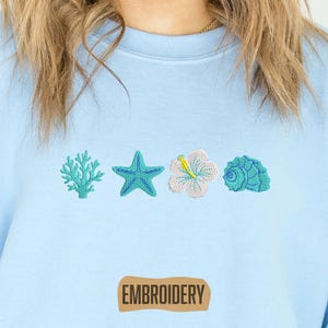 May include: Light blue sweatshirt with embroidered sea-themed designs: coral, starfish, hibiscus flower, and seashell. The word "EMBROIDERY" is printed on a brown rectangle at the bottom. The sweatshirt is a soft, casual garment.