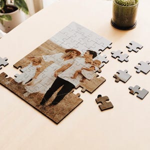 May include: A personalized family photo puzzle with pieces scattered on a light wood surface. The puzzle features a family portrait with a couple and two children. Several gray puzzle pieces are scattered around the partially assembled puzzle.