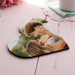May include: A heart-shaped jigsaw puzzle featuring a photograph of a couple embracing. The puzzle is made of multiple pieces and sits on a pink wooden surface. A white mug and flowers are in the background.