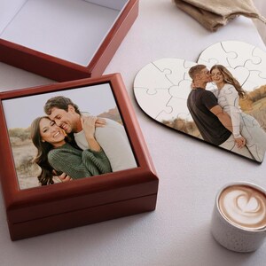 May include: A wooden box with a framed photo of a couple embracing, a heart-shaped puzzle with a photo of a couple, and a cup of coffee with latte art. The box is dark red, and the puzzle is made of wood.