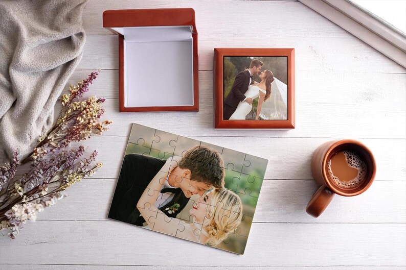 May include: A wedding-themed still life featuring a framed photo, a jigsaw puzzle, and a gift box, all with images of a couple. A mug of coffee and dried flowers are also present. The items are arranged on a white wooden surface.