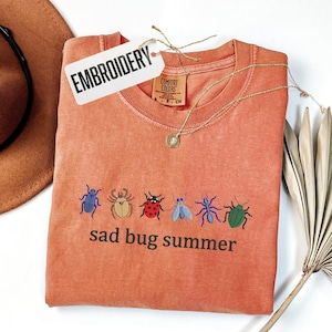 May include: An orange-red t-shirt with embroidered bugs and the text "sad bug summer." The shirt has a tag that says "EMBROIDERY." A gold necklace and a brown hat are also in the image.
