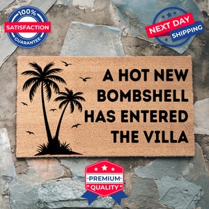 May include: A rectangular tan coir doormat featuring a black silhouette of two palm trees and several birds. The text printed in black reads: A HOT NEW BOMBSHELL HAS ENTERED THE VILLA. The mat has a coarse, natural fiber texture and a clean, straight edge.