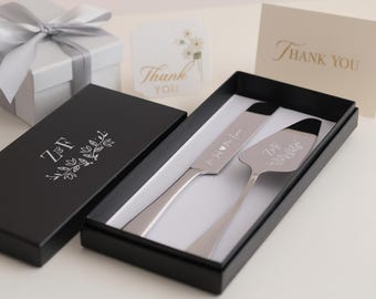 May include: A silver cake knife and server set in a black box with white interior. The knife and server are engraved with text and initials. A white gift box with a silver ribbon and a "Thank You" card are also in the image.