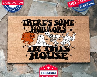 May include: A brown coir doormat with the text "There's some horrors in this house" in black. The text is in a retro font. There are illustrations of a ghost, a skeleton, a mummy, and a pumpkin with a scary face.
