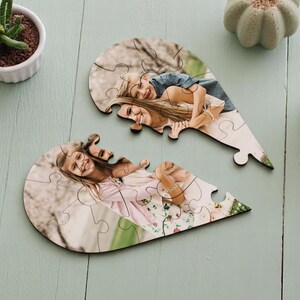 May include: Two heart-shaped jigsaw puzzles, each depicting a photograph of two people. The puzzles are on a light blue wooden surface, with a cup of coffee and a small potted plant nearby. The puzzles are made of wood.
