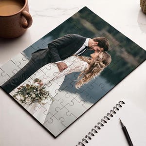 May include: A completed jigsaw puzzle featuring a romantic image of a couple embracing. The puzzle shows a man in a suit and a woman in a wedding dress. The puzzle is on a white surface with a cup of coffee and a plant.