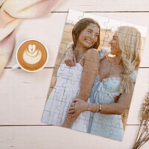 May include: A completed jigsaw puzzle featuring a photograph of two women embracing, set against a natural outdoor backdrop. The puzzle is on a white wooden surface, accompanied by a cup of coffee and a decorative scarf.