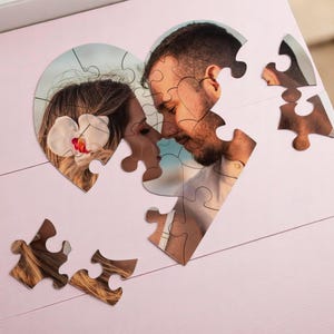 May include: A heart-shaped photo puzzle featuring a couple, with pieces scattered on a pink surface. The image shows a man and woman in a close embrace. A small potted cactus is in the upper left corner.
