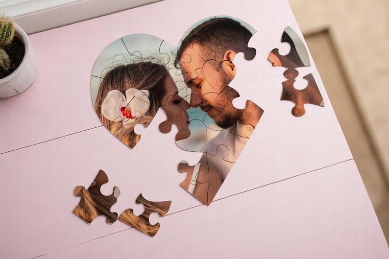 May include: A heart-shaped photo puzzle featuring a couple, with pieces scattered on a pink surface. The image shows a man and woman in a close embrace. A small potted cactus is in the upper left corner.