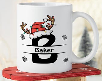 May include: White ceramic mug with a black letter "B" decorated with a reindeer wearing a Santa hat with Christmas lights. The name "Baker" is written below the letter.