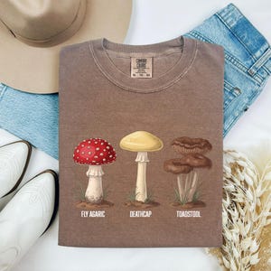 May include: A brown t-shirt featuring a graphic print of three mushrooms with the text "FLY AGARIC", "DEATHCAP", and "TOADSTOOL" printed underneath each respective illustration. The shirt has a crew neckline and a Comfort Colors brand label.