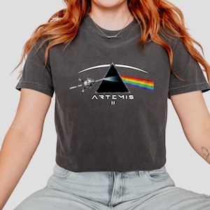 May include: Dark gray t-shirt featuring a graphic design with a black triangle, a rainbow, and a spacecraft. The text "ARTEMIS II" is printed below the triangle. The shirt has short sleeves and a crew neck.
