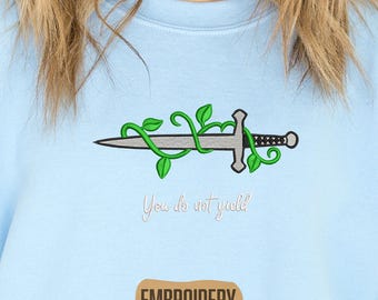 Puede incluir: Sudadera azul claro con un diseño de espada bordada entrelazada con vides verdes. El texto "You do not yield" está bordado debajo de la espada. La palabra "EMBROIDERY" está impresa en un rectángulo marrón en la parte inferior.