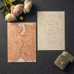 May include: A wedding invitation set featuring a laser-cut peach-colored floral cover, a cream-colored insert with printed text, and a gold ring in a velvet box. The text reads: Together with their families, Olivia and Richard invite you to their wedding, Saturday 14, 2027, at five o'clock in the evening, 123 anywhere st., any city, reception to follow.