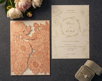 May include: A wedding invitation set featuring a laser-cut peach-colored floral cover, a cream-colored insert with printed text, and a gold ring in a velvet box. The text reads: Together with their families, Olivia and Richard invite you to their wedding, Saturday 14, 2027, at five o'clock in the evening, 123 anywhere st., any city, reception to follow.
