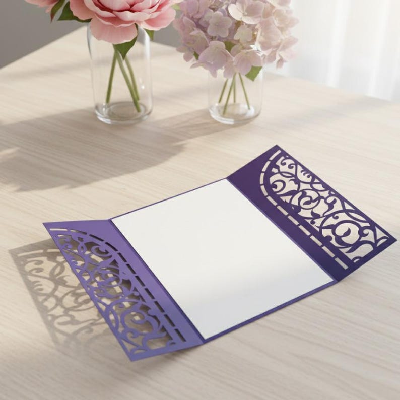 May include: A purple invitation card with intricate laser-cut designs, open to reveal a blank white space for text. The card is on a light wooden surface, with soft shadows. Two glass vases with flowers are in the background.