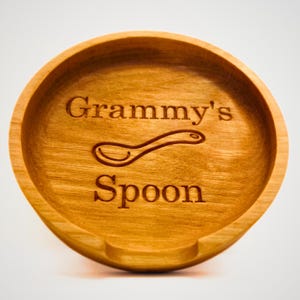 May include: A wooden spoon rest with the text "Grammy's Spoon" engraved on it. The spoon rest is made of light brown wood and has a cutout for a spoon.