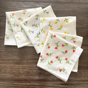 Puede incluir: Seis servilletas de tela blancas con un estampado floral. Cada servilleta presenta flores pequeñas de diferentes colores, incluyendo naranja, azul, amarillo y rojo, con tallos y hojas verdes. Las servilletas están dispuestas sobre una superficie de madera oscura.