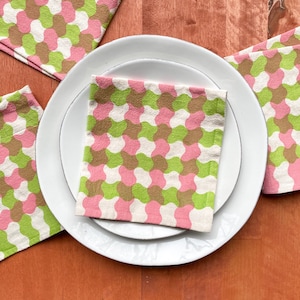 May include: A set of four pink, green, and brown patterned fabric napkins on a white plate. The napkins have a geometric design.