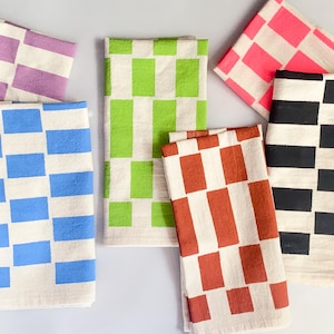 May include: A collection of colorful checkered pattern towels. The towels feature a repeating pattern of rectangles in various colors, including purple, blue, green, red, pink, and black, set against a neutral background. The towels are neatly folded and arranged.