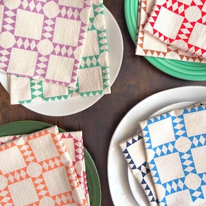 May include: A selection of patterned cloth napkins in shades of purple, green, red, orange, and blue. Each napkin displays a geometric design with squares, diamonds, and circles. The napkins are arranged on white and green plates, creating a colourful table setting.