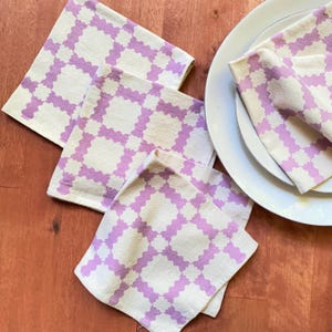 Puede incluir: Cuatro servilletas de tela con estampado morado y blanco dobladas sobre una superficie de madera. Una servilleta está doblada sobre un plato blanco.