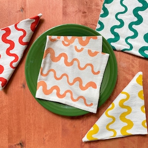 May include: Four fabric napkins with a wavy pattern. The napkins are in different colours: red, orange, green, and yellow. The napkins are on a wooden surface.