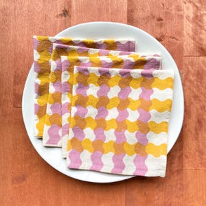 Può includere: Quattro tovaglioli di stoffa rosa e gialli piegati e impilati su un piatto bianco. I tovaglioli hanno un motivo geometrico con forme arrotondate.