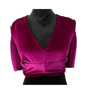 May include: A magenta velvet crop top with a deep V-neck and short sleeves. The top is displayed on a black mannequin, showcasing its design and texture. The fabric appears soft and luxurious, with a gathered detail at the waist.