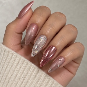 May include: Almond-shaped nails featuring a mix of finishes including metallic rose gold, shimmering pink, a glittery accent nail with star-shaped sequins, and a pink nail adorned with three small clear rhinestones.