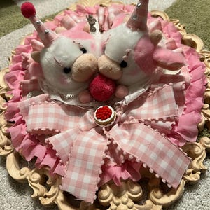 May include: A decorative wall hanging featuring two plush cow figures with pink and white fur, wearing party hats. The design includes a pink gingham bow, ruffled trim, and a small cake embellishment, set within a carved wooden frame.