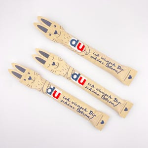Könnte beinhalten: Drei braune Papierpäckchen mit Hasenohren und dem Text "du" in Rot und Blau. Jedes Päckchen hat den Text "ich wunsch Dip schöne Ostern!" aufgedruckt.