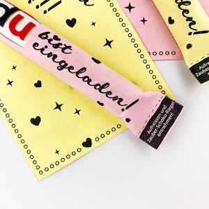 May include: Pink and yellow paper wrappers with black text that reads "Du bist eingeladen!" which translates to "You are invited!" The wrappers are decorated with hearts and stars. The text on the bottom of the wrapper reads "Aufreissen und Zauber-Schoko-Riegel entnehmen!" which translates to "Tear open and remove the magic chocolate bar!"