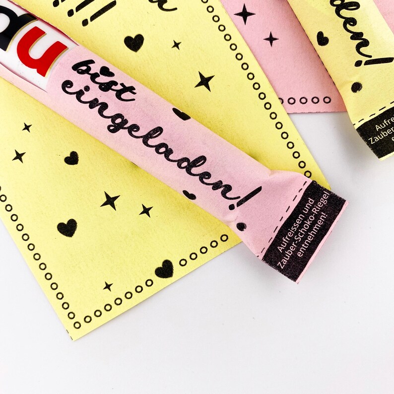 May include: Pink and yellow paper wrappers with black text that reads "Du bist eingeladen!" which translates to "You are invited!" The wrappers are decorated with hearts and stars. The text on the bottom of the wrapper reads "Aufreissen und Zauber-Schoko-Riegel entnehmen!" which translates to "Tear open and remove the magic chocolate bar!"