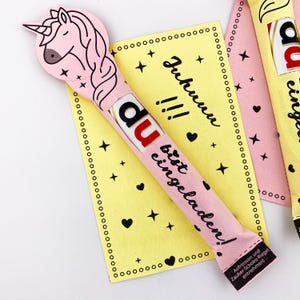 May include: A pink and yellow card with the text "Juhuuu!!! du bist eingeladen!" and a pink chocolate bar with a unicorn design and the text "du" on it.