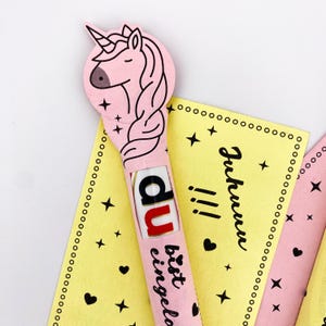 May include: A pink unicorn-shaped card with a chocolate bar inside. The card has the text "Du bist eingeladen" and stars and hearts printed on it.