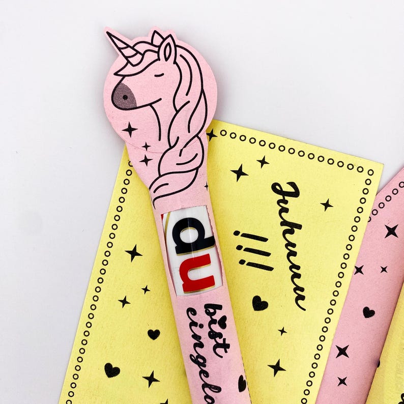 May include: A pink unicorn-shaped card with a chocolate bar inside. The card has the text "Du bist eingeladen" and stars and hearts printed on it.