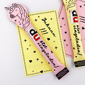May include: A pink and yellow card with a unicorn design and the text "Du bist eingeladen!"  The card is decorated with black stars and hearts. The text "Juhunu!!!" is written on the card.