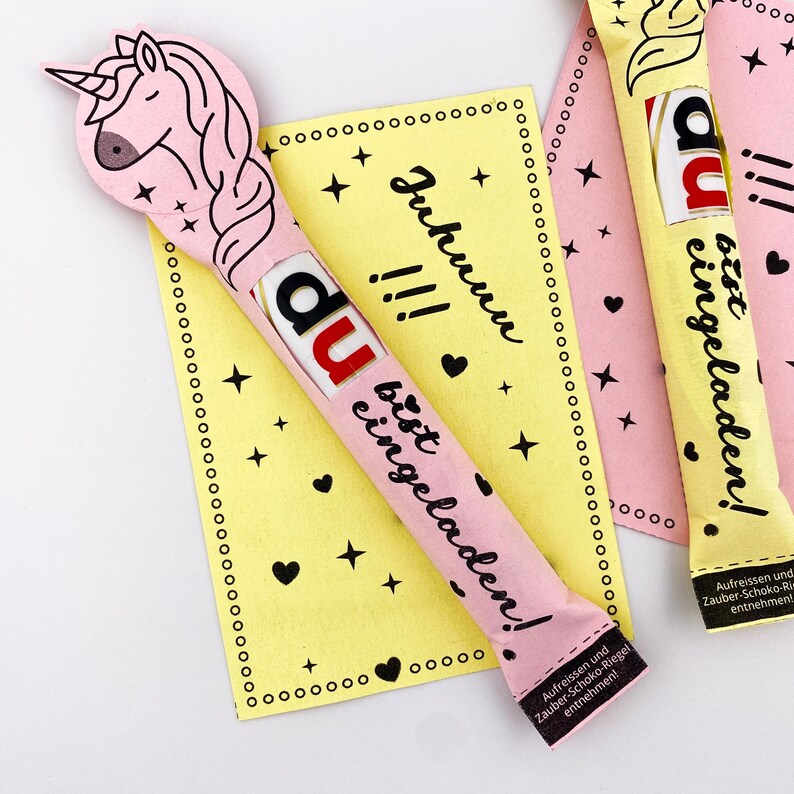 May include: A pink and yellow card with a unicorn design and the text "Du bist eingeladen!"  The card is decorated with black stars and hearts. The text "Juhunu!!!" is written on the card.