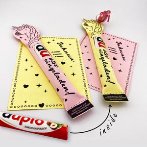 May include: Two pink and yellow unicorn-shaped chocolate bar wrappers with the text "Juhunu !!! du bist eingeladen!"  The wrappers are folded and ready to be filled with a chocolate bar.