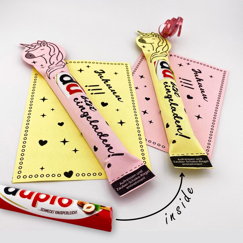 May include: Two pink and yellow unicorn-shaped chocolate bar wrappers with the text "Juhunu !!! du bist eingeladen!"  The wrappers are folded and ready to be filled with a chocolate bar.