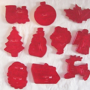 May include: A collection of ten red plastic cookie cutters in various shapes including a train, a round ornament, a maple leaf, a Christmas tree, a snowman, a Santa Claus figure, an angel, a wreath, a rectangular gift, and a reindeer.