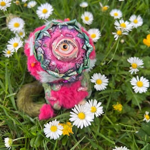 Puede incluir: Un juguete de peluche hecho a mano de color rosa y verde con un ojo grande y una corona de hojas. El juguete está sentado en un campo de margaritas blancas.