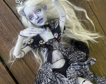 May include: A detailed art doll with pale skin, long white hair, and a black and white patterned dress. The doll has a crown and is posed with arms outstretched. The doll's face has purple and blue accents, and it is lying on a wooden surface.